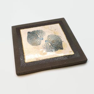 Op de afbeelding: Een vierkante keramische onderzetter met een donkerbruine rand en een crèmekleurig midden. Het midden heeft een blauw bladerontwerp met gouden accenten. De onderzetter is een decoratief item voor thuisgebruik.