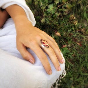 May include: A silver ring with a large, oval-shaped red and gold stone. The stone has a textured surface with a gold crisscross pattern. The ring is on a hand, with a white fabric in the background.