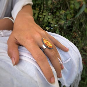 May include: A silver ring with an oval-shaped, textured, orange and gold design. The ring is worn on a finger, with a white fabric and green foliage in the background. The ring's design has a dark border.