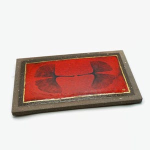 Può includere: Vassoio rettangolare in ceramica con un centro rosso con un disegno di foglie nere. Il vassoio ha un bordo dorato e una cornice grigio scuro. Il design è simmetrico, con due foglie contrapposte.