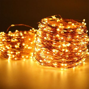May include: Two bundles of warm white LED string lights. The lights are on a thin copper wire, coiled into a circular shape. The background is a dark, warm tone, highlighting the glowing lights. These lights are ideal for home decor.