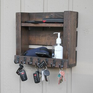 May include: A dark brown wooden wall mounted key rack with hooks and a shelf. The shelf has a small bottle of hand sanitizer and a black phone case on it. There are several keys hanging from the hooks.