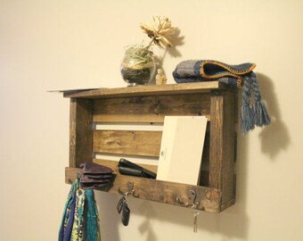 May include: A rustic wooden shelf with a key hook and a small shelf for storage. The shelf is stained a dark brown and has a natural wood finish. It is mounted on a white wall and has a blue and white scarf hanging from the hooks.