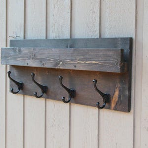 Pu&ograve; includere: Un appendiabiti in legno rustico con uno scaffale e quattro ganci in metallo nero. L'appendiabiti &egrave; tinto di un colore marrone scuro e ha una finitura invecchiata.