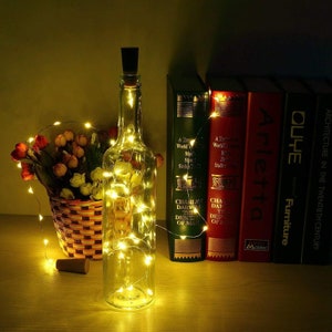 May include: A clear glass bottle with a cork stopper is wrapped in a string of warm white fairy lights. The bottle is sitting on a wooden surface in front of a bookshelf with several books. The books have titles like "A Treasury of the World's Most Famous Stamps" and "Arletta Furniture".