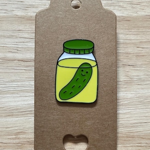 Pode incluir: Um broche de esmalte de um picles num frasco. O broche apresenta um picles verde com um rosto sorridente dentro de um frasco de vidro com uma tampa verde. O frasco é preenchido com líquido amarelo. O broche está preso a uma etiqueta castanha com um recorte em forma de coração.