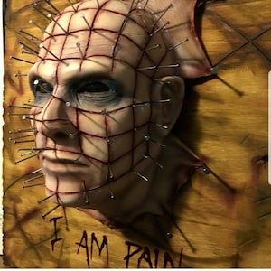 May include: A 3D sculpted face mask with a grid of nails and red lines, mounted on a wooden surface. The mask is painted to look like skin and has a realistic expression. The text "I AM PAIN" is written below the mask.