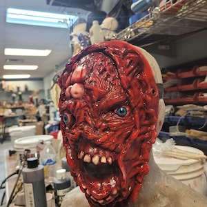 Puede incluir: Una máscara de terror de látex roja y blanca con una apariencia grotesca y carnosa. La máscara tiene la boca abierta de par en par con dientes y un solo ojo visible.