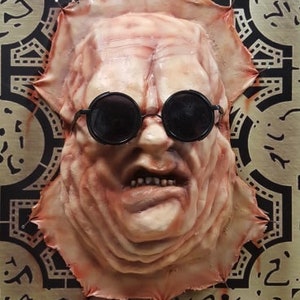 May include: A close-up of a fleshy, pink, and white mask with black sunglasses. The mask has a detailed, textured surface and a slightly open mouth. The mask is displayed on a black and white background with a geometric pattern.