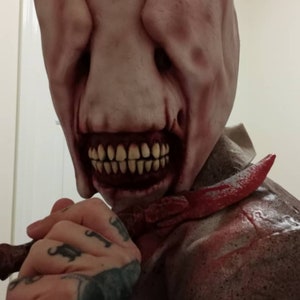 Może przedstawiać: Zbliżenie na osobę noszącą jasnoróżową i czerwoną maskę horroru z szerokim, zębatym uśmiechem. Maska ma duży, mięsny nos i wydaje się być wykonana z lateksu. Osoba trzyma czerwony nóż w ręku.