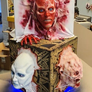 Puede incluir: Una caja en forma de cubo con un diseño de patrón dorado y negro. Tres caras de la caja tienen una cabeza humana que sobresale de ellas. Las cabezas están hechas de un material similar a la carne y tienen rasgos realistas. La parte superior de la caja tiene una tela blanca drapeada sobre ella, con una cabeza roja que sobresale de la tela. La cabeza también está hecha de un material similar a la carne y tiene rasgos realistas.