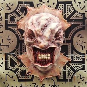 May include: A grotesque, fleshy mask with a wide, toothy grin and a textured, veiny appearance. The mask is mounted on a wooden panel with a geometric pattern and is secured with wire. The overall aesthetic is horror-themed.