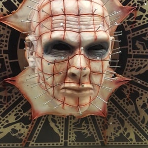 May include: A close-up of a horror movie prop mask. The mask is a human face with a grid of red lines and many small silver pins sticking out. The mask is made of a light-colored material and has a realistic appearance.