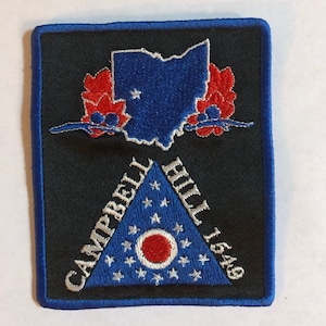 Puede incluir: Un parche bordado negro con un borde azul. El parche presenta un contorno azul del estado de Ohio con una estrella blanca en el centro. Debajo del contorno del estado hay un triángulo azul con estrellas blancas y un círculo rojo en el centro. El texto "Campbell Hill 1549" está bordado en blanco en el triángulo.