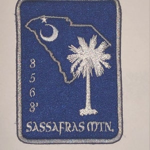 May include: A blue embroidered patch with a white outline. The patch features a white outline of the state of South Carolina with a crescent moon and a palmetto tree. The text "SASSAFRAS MTN." is embroidered in white at the bottom of the patch.