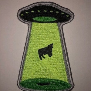 Könnte beinhalten: Ein gestickter Aufnäher, der ein UFO mit einer schwarzen Kuh im Lichtstrahl zeigt, in Grün und Grau.