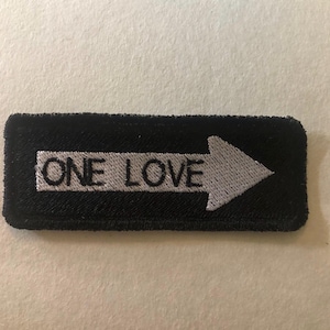 Könnte beinhalten: Schwarzer rechteckiger Aufnäher mit den weißen Worten "ONE LOVE" und einem weißen Pfeil nach rechts. Der Aufnäher ist gestickt und hat eine strukturierte Optik.