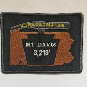 Può includere: Toppa rettangolare nera con testo e disegno ricamati. La toppa presenta le parole "A GEOLOGIC FEATURE" sopra un contorno marrone di una montagna e il testo "MT. DAVIS 979m". Una piccola stella è ricamata in basso a sinistra.