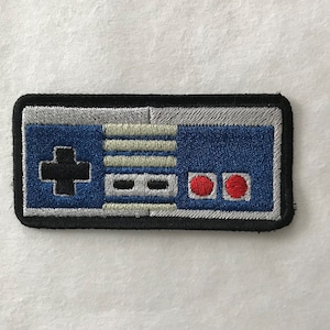 May include: A black and blue embroidered patch of a classic video game controller. The patch features a directional pad, four buttons, and two red buttons.