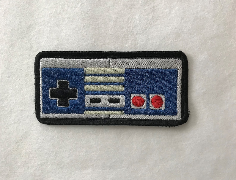 Peut inclure: Un patch brod&eacute; noir et bleu repr&eacute;sentant une manette de jeu vid&eacute;o classique. Le patch pr&eacute;sente un pav&eacute; directionnel, quatre boutons et deux boutons rouges.