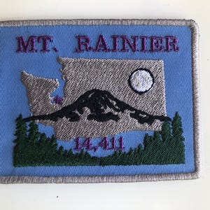 Puede incluir: Parche bordado con el Monte Rainier, el contorno del estado de Washington y el texto "MT. RAINIER 14.411". El diseño incluye una montaña negra, una luna blanca y árboles verdes, todo sobre un fondo azul.