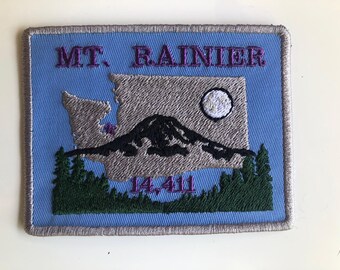 May include: Embroidered patch featuring Mt. Rainier, the Washington state outline, and the text "MT. RAINIER 14,411". The design includes a black mountain, a white moon, and green trees, all on a blue background.