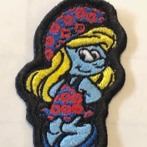 Pode incluir: Um remendo bordado com um personagem azul parecido com um Smurf com cabelo amarelo, usando um chapéu e um maiô com estampa vermelha e azul. O personagem está sorrindo e tem um olho branco.