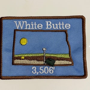 Puede incluir: Un parche bordado azul con un borde marrón. El parche presenta una imagen estilizada de un paisaje con un sol amarillo, un campo verde, una colina marrón y un poste blanco con un objeto verde en la parte superior. El texto "White Butte" está en la parte superior del parche y "3,506'" en la parte inferior.