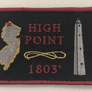 May include: A black embroidered patch with red stitching around the edges. It features a gray outline of the state of New Jersey with a red star on top, the words "HIGH POINT" in red, a gold infinity symbol, and the number "1803'" in red. A gray obelisk is on the right side of the patch.