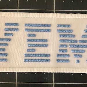 Pode incluir: Patch bordado com as letras "IBM" em azul claro sobre um fundo retangular branco. O patch tem uma borda branca e é projetado para ser costurado ou passado a ferro no tecido. Ideal para entusiastas de tecnologia.