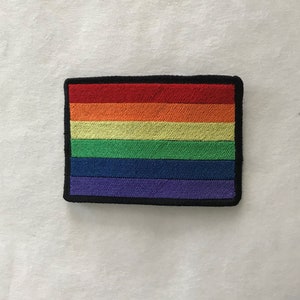 Puede incluir: Un parche de bandera del orgullo arcoíris con seis rayas horizontales en los colores rojo, naranja, amarillo, verde, azul y morado. El parche está rodeado por un borde negro.