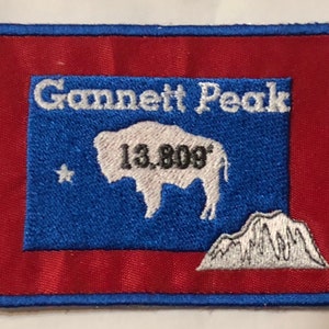 Op de afbeelding: Een geborduurde rode en blauwe patch met de tekst "Gannett Peak 13,808'" en een wit silhouet van een buffel. De patch heeft een wit bergketen in de rechterbenedenhoek.