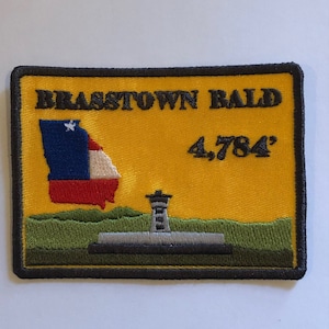 Puede incluir: Un parche bordado amarillo con borde negro. El parche presenta un contorno estilizado del estado de Georgia con una estrella en la esquina superior izquierda. El texto "Brasstown Bald 4,784'" está bordado en negro. El parche también presenta una torre estilizada con un fondo verde.