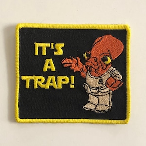Puede incluir: Un parche bordado negro y amarillo con un personaje de dibujos animados y el texto "It's a Trap!"