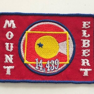Könnte beinhalten: Roter gestickter Aufnäher mit weißem Rand. Der Aufnäher zeigt einen weißen Kreis mit rotem und weißem Rand um einen gelben Kreis mit einem weißen Stern. Der Text "MOUNT ELBERT 14,439" ist in Weiß gestickt.