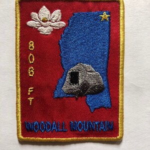 Puede incluir: Un parche bordado con un borde dorado. El parche presenta un contorno azul del estado de Mississippi con una flor de magnolia blanca y una estrella dorada. El texto "Woodall Mountain" y "800 ft" también está bordado en el parche.