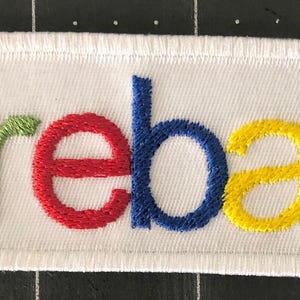 May include: Embroidered white patch with the word "reba" in colourful letters: green, red, blue, and yellow. The patch has a white border and is rectangular in shape.