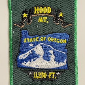 Op de afbeelding: Een groene geborduurde patch met een gele rand. De patch toont een gestileerde afbeelding van Mount Hood, Oregon, met de tekst "HOOD MT." en "STATE OF OREGON" boven de berg en "11,250 FT." eronder. Twee gele sterren bevinden zich aan weerszijden van de tekst "HOOD MT."