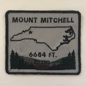 Pode incluir: Um remendo bordado cinza com costuras pretas que representa o contorno da Carolina do Norte com o texto "MOUNT MITCHELL 6684 FT."