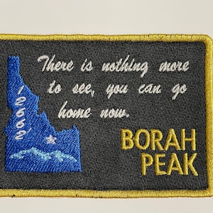 Puede incluir: Parche bordado con fondo negro y ribete dorado. El parche presenta el texto "There is nothing more to see, you can go home now." y "BORAH PEAK". También se incluye un contorno azul de Idaho con el número "12662".