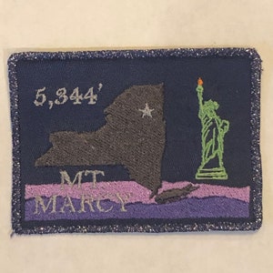 Puede incluir: Un parche bordado azul con un contorno gris del estado de Nueva York, una Estatua de la Libertad verde y el texto "5,344' MT MARCY".
