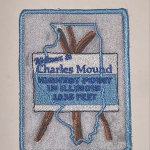 Op de afbeelding: Een blauwe en witte geborduurde patch met de tekst "Welcome to Charles Mound" en een omtrek van de staat Illinois. De patch is op een witte achtergrond en heeft een blauwe rand.