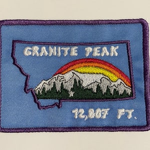 Puede incluir: Un parche bordado azul con un contorno blanco que representa el estado de Montana con un arcoíris sobre una cordillera. El texto "GRANITE PEAK 12,867 FT." está bordado en blanco.