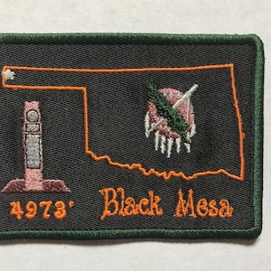 May include: An embroidered patch with an orange outline of the state of Oklahoma. The patch features a pink and white building, a Native American headdress, and the text "Black Mesa 4973'" in orange.