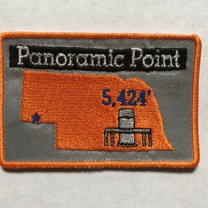 Puede incluir: Un parche bordado gris y naranja con el texto "Panoramic Point" y "5,424'" en blanco. El parche presenta un contorno estilizado de un estado con una torre en el centro.