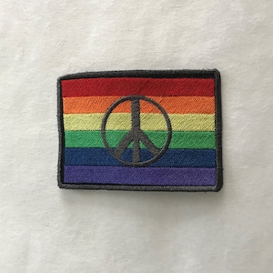 Może przedstawiać: Naszywka w kolorach tęczy z czarnym symbolem pokoju w środku. Naszywka jest otoczona czarną ramką.