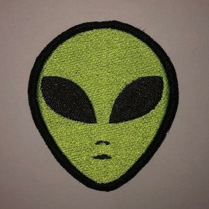 Könnte beinhalten: Grüne Alien-Gesichts-Stickerei mit schwarzen Augen und schwarzem Umriss.