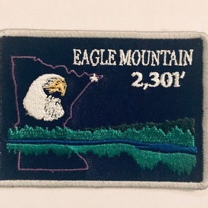 May include: A blue and green embroidered patch with a white outline. The patch features a white eagle head, a star, and the text "Eagle Mountain 2,301'" in white.