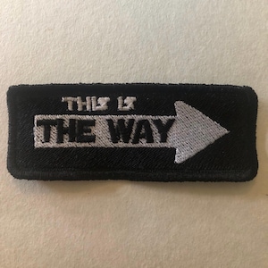 May include: Black rectangular patch with white embroidered text and arrow. The text reads "THIS IS THE WAY". The arrow points to the right. The patch is likely for clothing or accessories.