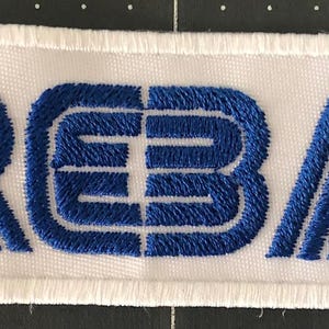 May include: Embroidered patch featuring the letters "RBA" in blue on a white rectangular background. The letters are in a bold, stylized font, with a white border.